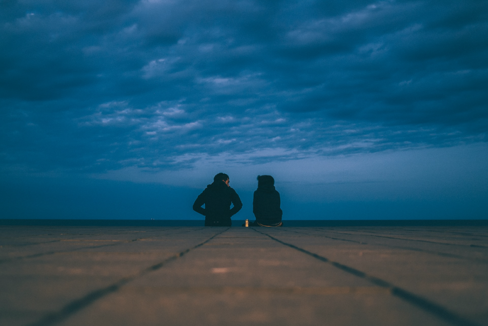 Image: from a worm's eye view, a couple wearing dark hoodies are seen sitting at the edge of a brick sea wall, turned to face the open ocean, under a heavily-clouded sky at early evening.