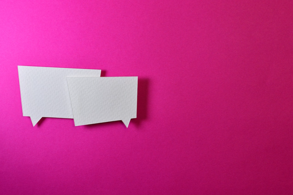 Image: two overlapping cartoon-style word balloons made from cut white paper sit atop a hot pink background
