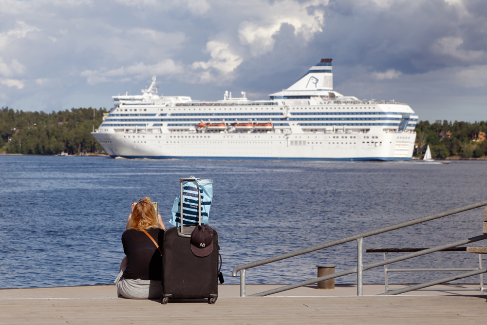 Image: a woman sits on a pier with her luggage, watching the cruise ship which has sailed away without her.