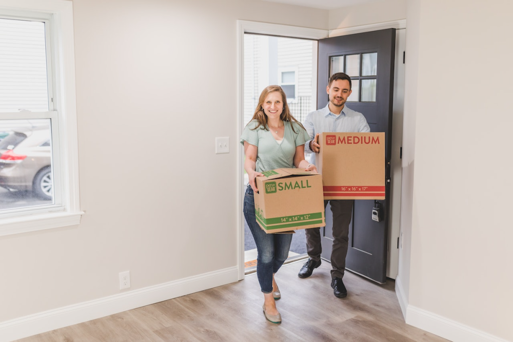Image: a couple carries moving boxes through the front door of their empty new home.