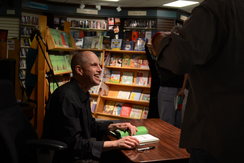 Image: An author smiles at an audience member as he prepares to autograph a book at an in-store book signing.