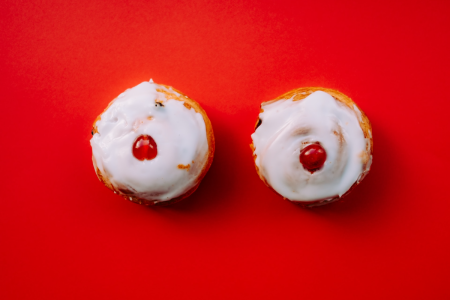 Image: on a red tabletop are two cinnamon rolls side by side, each topped with white icing and a sliced maraschino cherry, suggestively resembling a pair of breasts.