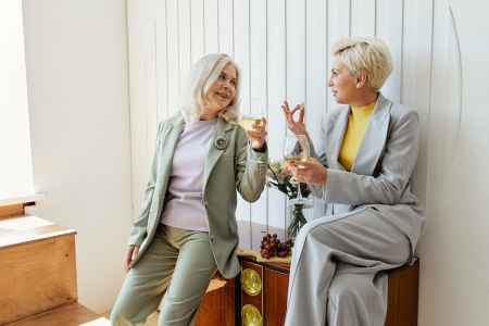 Image: two women in business-casual attire hold a conversation while enjoying wine and cheese in a room with elegant modern decor.