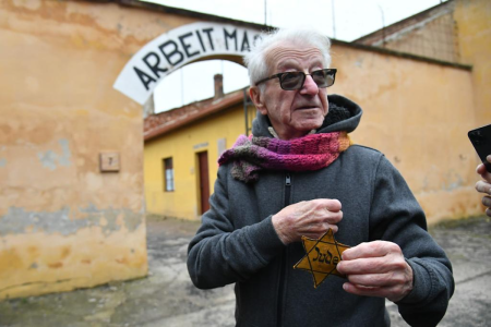 Image: Holocaust survivor Gidon Lev stands at the gate of the former Terezin concentration camp in the Czech Republic, holding a yellow fabric Magen David with the word "Jude" stitched on it.