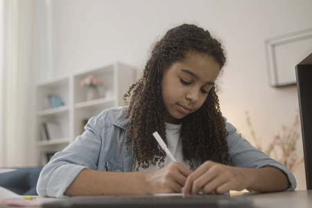 Image: a girl in her early teens sits writing at a table.
